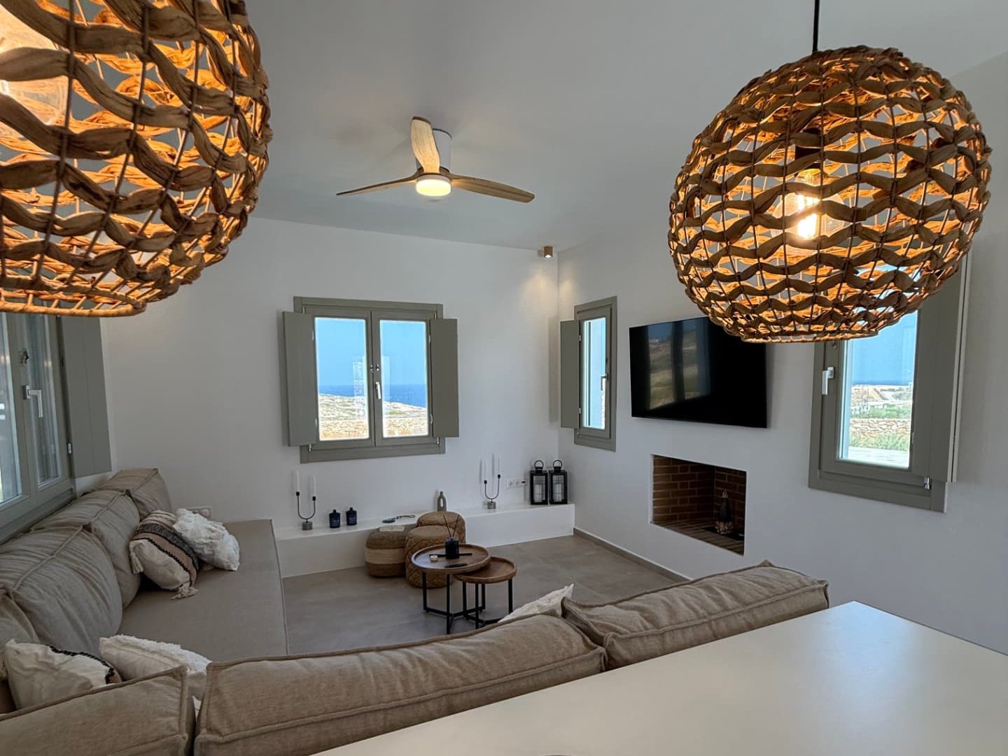 The living room at DEON, woven pendants above a long linen sofa with the Aegean framed in the shuttered windows