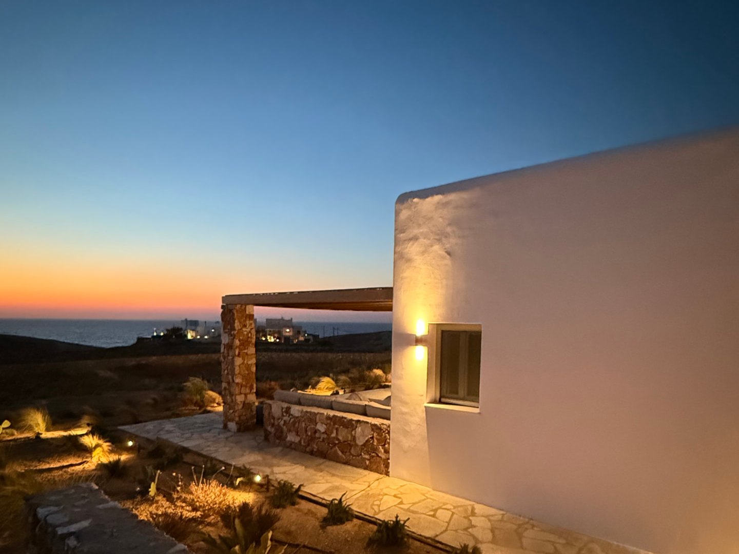 The whitewashed volume and stone pergola at DEON against an orange sunset over the Aegean